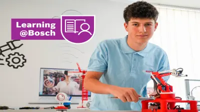 Un grup de oameni stau la o masă râzând și lucrează împreună la diferite sarcini în cadrul hackathon-ului "Bosch Connected Experience" din Berlin. Masa este plină cu diferite bilețele, foi colorate, creioane și un laptop. În spate, alți participanți stau în jurul unui panou cu diferite postere de proiecte.